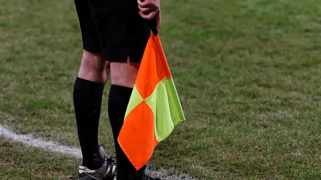 Un árbitro asistente sostiene su banderín naranja y amarillo junto a la línea de banda en un partido de futbol nocturno