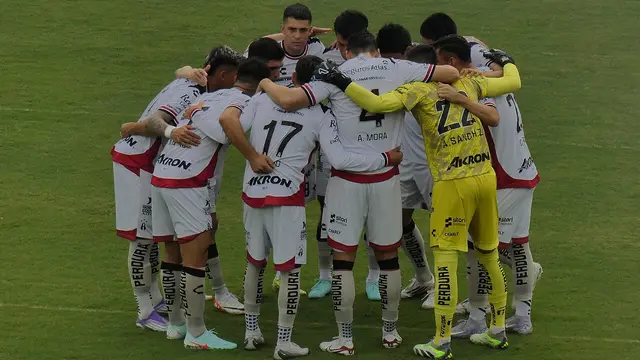 Jugadores del Atlas abrazados en círculo sobre la cancha antes de un partido, con su uniforme blanco con detalles rojos y el portero de amarillo