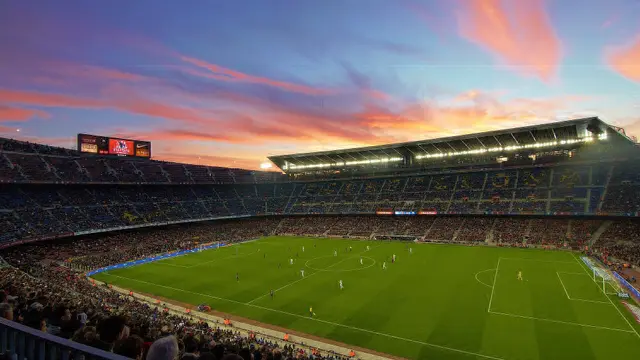 El Camp Nou de Barcelona lleno durante un partido, con la cancha iluminada y un cielo rosa y naranja al atardecer