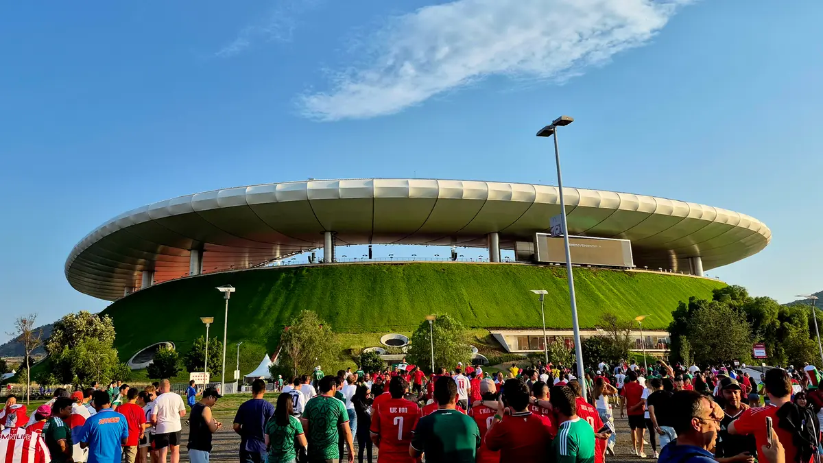 Aficionados caminan frente al Estadio Akron, casa de Chivas en Zapopan, con su techo blanco y su colina de pasto verde en una tarde soleada