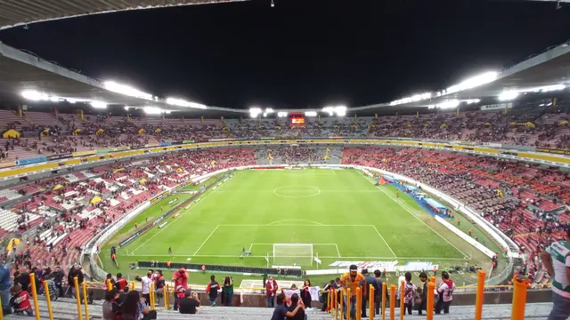 Interior del Estadio Jalisco de noche, con la cancha iluminada y aficionados en las gradas vistos desde detrás de la portería sur