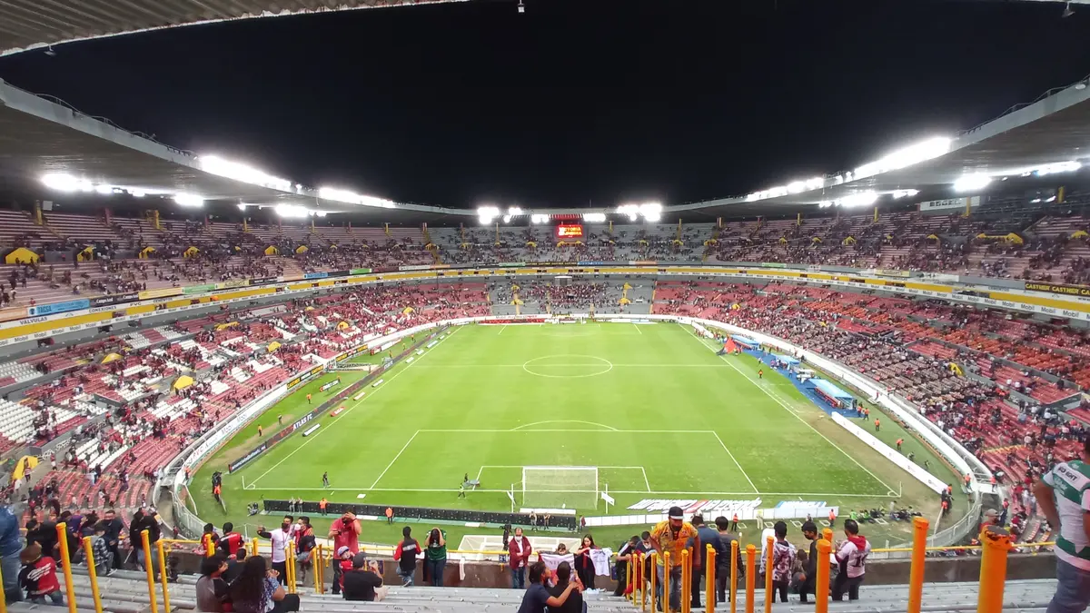 Interior del Estadio Jalisco de noche, con la cancha iluminada y aficionados en las gradas vistos desde detrás de la portería sur