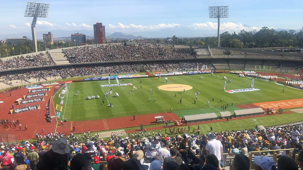 Tribunas llenas del Estadio Olímpico Universitario antes de un partido de futbol, con la cancha, la pista de atletismo y las torres de iluminación bajo un cielo despejado