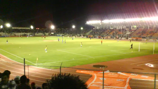 Partido nocturno en un estadio con pista de atletismo, gradas iluminadas y una gran playera naranja extendida junto a la cancha