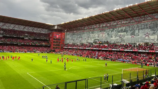 Jugadores de Toluca calientan en la cancha del Estadio Nemesio Díez, con las gradas llenas de aficionados de rojo y un gran mosaico en la tribuna