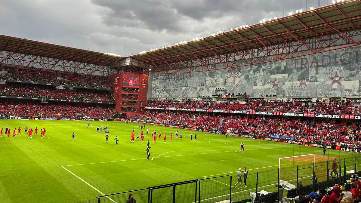 Jugadores de Toluca calientan en la cancha del Estadio Nemesio Díez, con las gradas llenas de aficionados de rojo y un gran mosaico en la tribuna