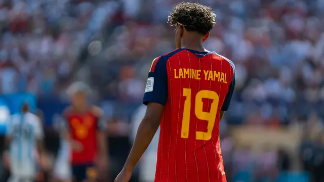 Lamine Yamal de espaldas con la camiseta roja número 19 de España durante un partido, con la tribuna llena al fondo
