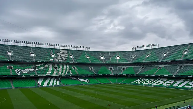 Interior del Estadio Benito Villamarín del Real Betis en Sevilla, con gradas verdes vacías y el cielo nublado
