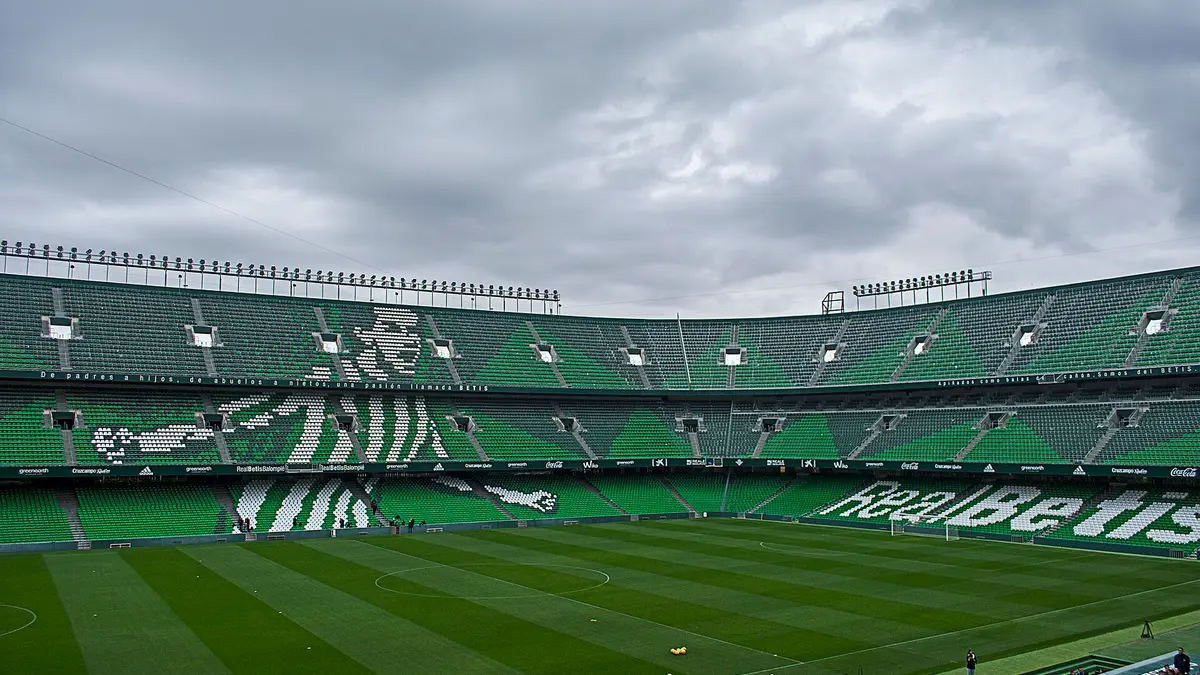 Interior del Estadio Benito Villamarín del Real Betis en Sevilla, con gradas verdes vacías y el cielo nublado