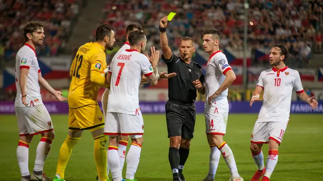 Un árbitro vestido de negro muestra la tarjeta amarilla mientras varios jugadores de blanco le reclaman en un partido de eliminatoria europea