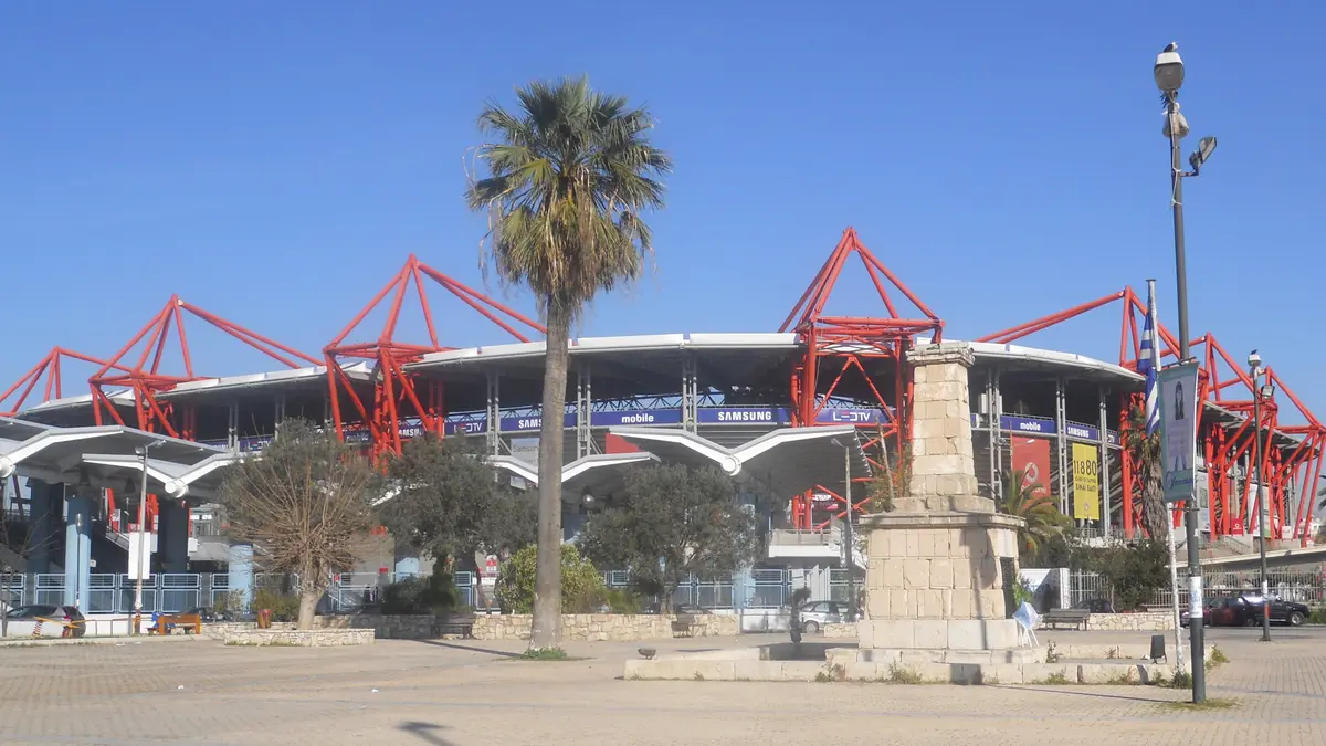 Exterior del Estadio Georgios Karaiskakis del Olympiacos en El Pireo, con su estructura roja, una palmera y un monumento de piedra bajo cielo azul