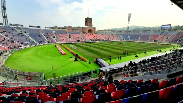 Interior del Stadio Renato Dall'Ara de Bolonia con la cancha, las gradas rojas y azules y la torre de ladrillo al fondo