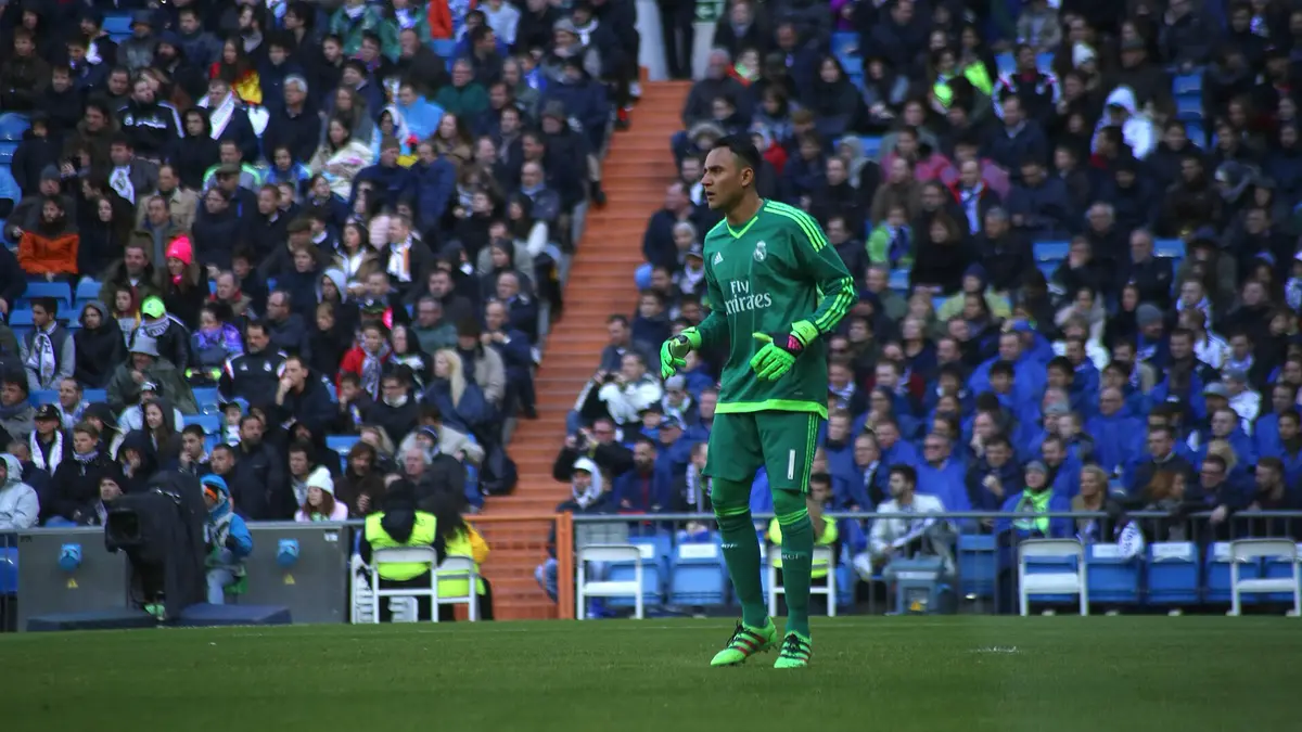Keylor Navas, con uniforme verde de portero y guantes, de pie en la cancha durante un partido con el Real Madrid, con las gradas llenas al fondo