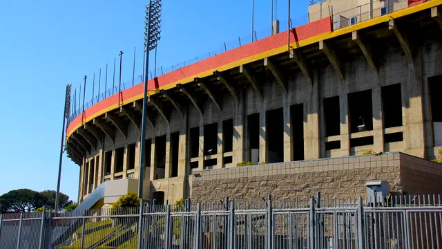 Exterior del Los Angeles Memorial Coliseum con sus torres de iluminación y una reja en primer plano bajo un cielo despejado