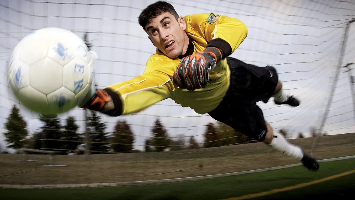 Portero con uniforme amarillo y guantes se lanza en el aire para atajar un balón frente a la red