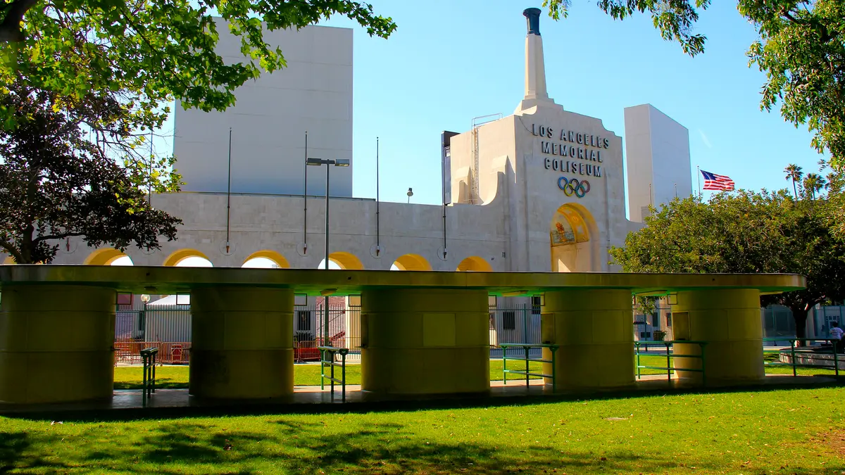 Peristilo del Los Angeles Memorial Coliseum con su torre, los aros olímpicos y la bandera de Estados Unidos, visto desde un jardín