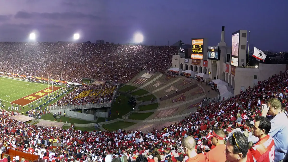 Gradas llenas del Los Angeles Memorial Coliseum al atardecer, con la cancha iluminada y el peristilo con el pebetero olímpico al fondo