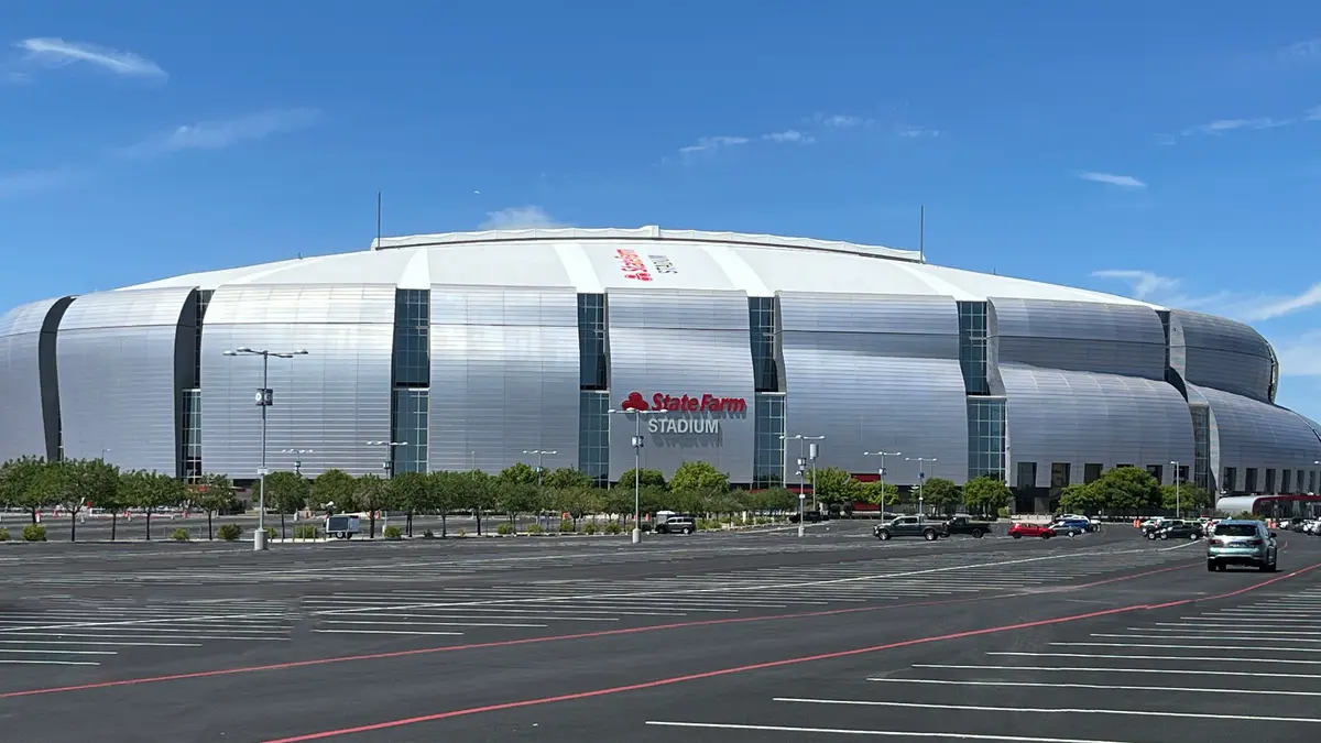 Exterior del State Farm Stadium en Glendale, Arizona, con su techo plateado y el estacionamiento vacío bajo cielo azul