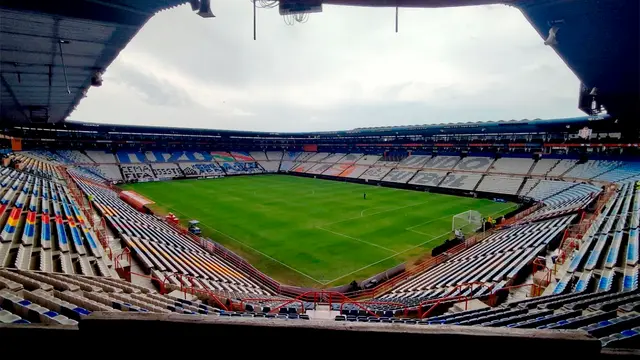 Vista desde la tribuna del Estadio Hidalgo vacío, con la cancha de pasto y las gradas de colores bajo un cielo nublado