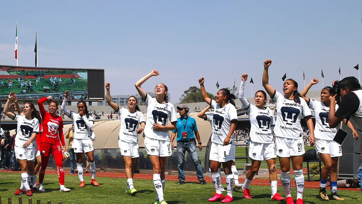 Jugadoras de Pumas Femenil con uniforme blanco levantan el puño frente a la tribuna del Estadio Olímpico Universitario en 2020