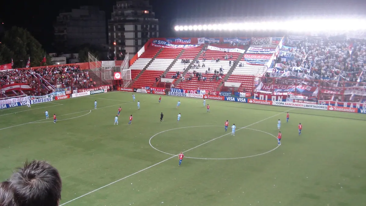 Partido nocturno en el Estadio Diego Armando Maradona de Argentinos Juniors, en Buenos Aires, con jugadores en la cancha y tribunas llenas de banderas