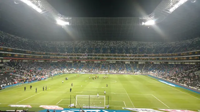 Cancha del Estadio BBVA iluminada de noche durante el calentamiento previo a un partido de Monterrey, con las gradas azules y blancas al fondo