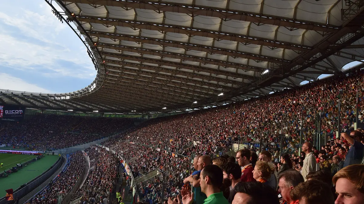 Tribunas llenas de aficionados en el Estadio Olímpico de Roma durante un partido de la AS Roma, con el techo del estadio iluminado por el sol