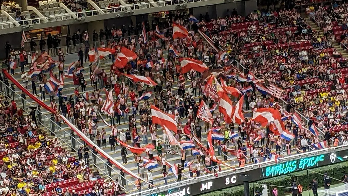 Aficionados de Chivas ondean banderas rojiblancas en una tribuna del Estadio Akron durante un Clásico ante el América, con seguidores azulcremas de amarillo abajo