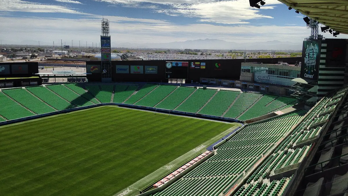 Interior del Estadio Corona de Torreón vacío, con la cancha de pasto y las gradas de butacas verdes bajo un cielo con nubes