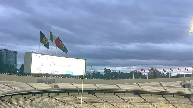 Gradas vacías del Estadio Olímpico Universitario bajo un cielo nublado y lluvioso, con el marcador electrónico y banderas al fondo