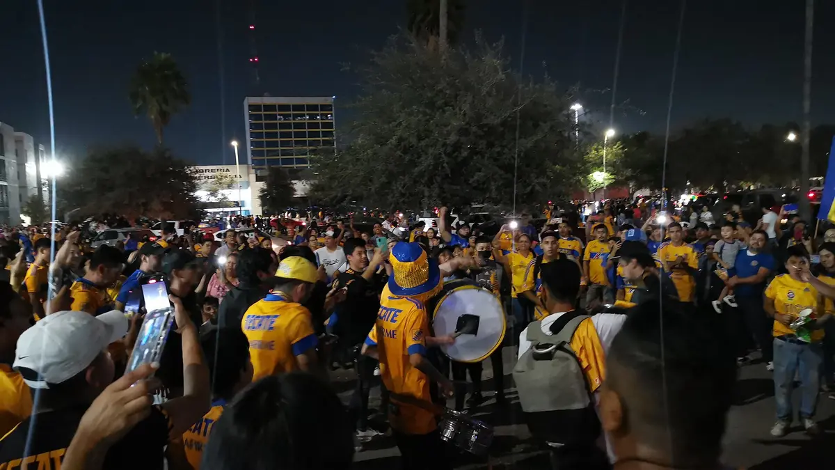 Aficionados de Tigres con playeras amarillas tocan tambores y celebran de noche en un estacionamiento antes de un partido de Tigres Femenil en 2022
