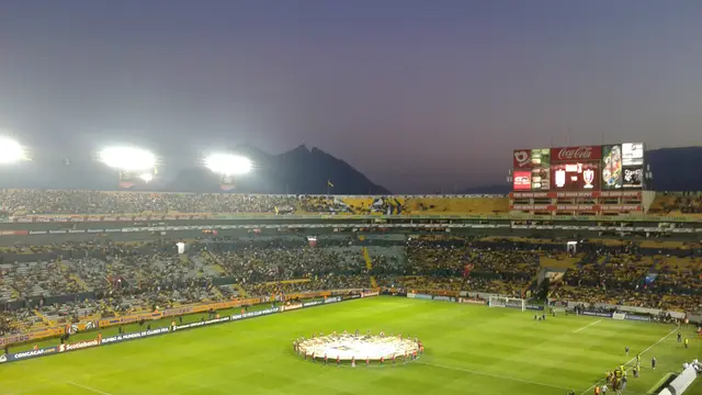 Estadio Universitario de Tigres lleno al atardecer, con el cerro al fondo y los jugadores alrededor del círculo central antes de un partido