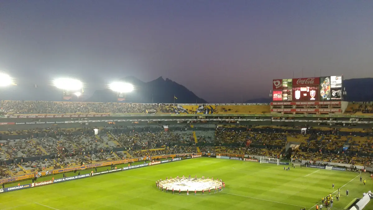 Estadio Universitario de Tigres lleno al atardecer, con el cerro al fondo y los jugadores alrededor del círculo central antes de un partido