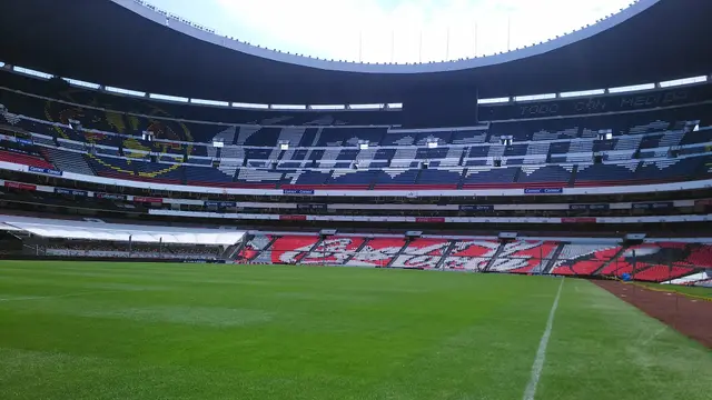 Cancha y tribunas vacías del Estadio Azteca vistas desde el nivel del césped, con las butacas de colores bajo el techo del estadio